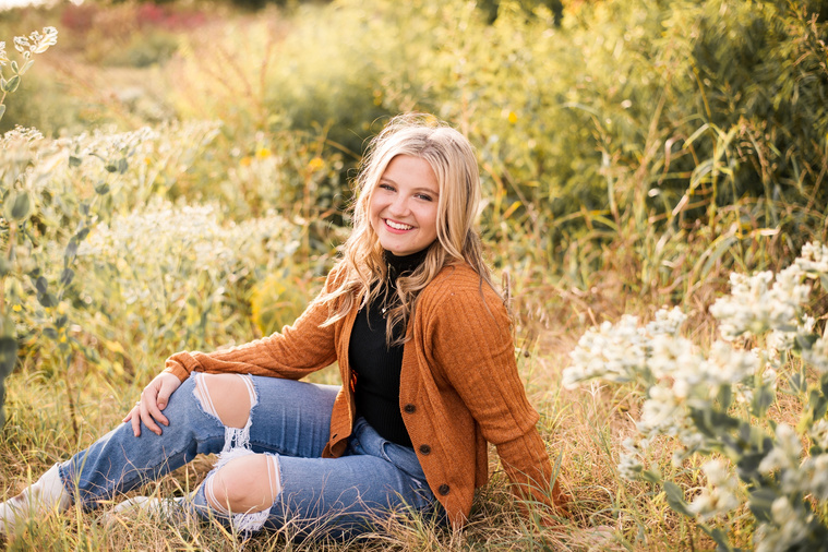 high school senior girl with blond hair wearing a rust colored sweater and jeans, sits in a golden field in Oklahoma smiling for her photo session