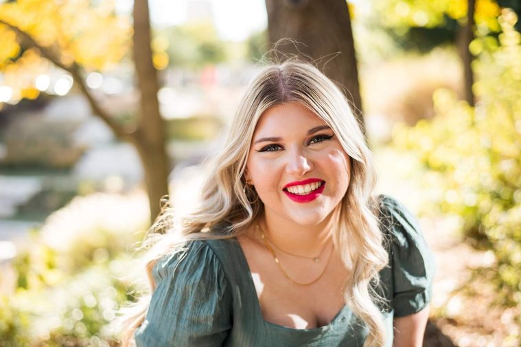 blond high school senior girl wearing a green dress smiles brightly at Myriad Gardens in downtown Oklahoma City for senior portraits