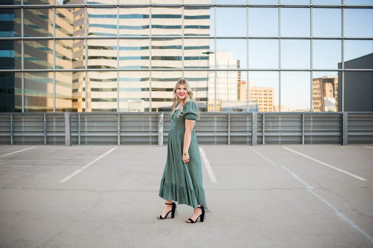 blond high school senior girl stands sideways looking at camera in a green dress on top of the devon tower parking garage in okc with reflective glass behind her