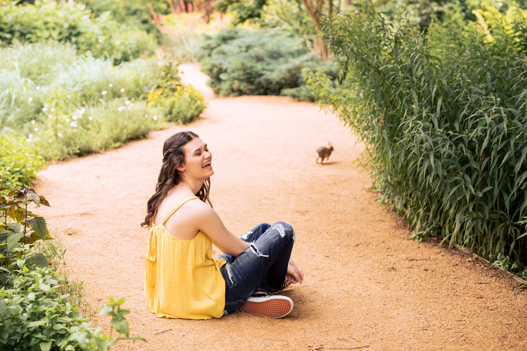 high school senior girl with long brown hair wearing a yellow top and jeans sits on a path laughing with a bunny hopping away at Myriad Gardens in Oklahoma City
