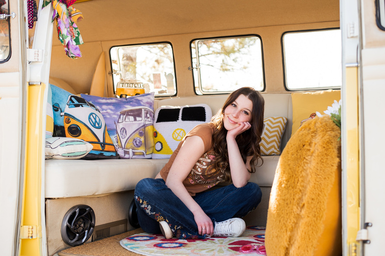 high school senior girl with brown hair wearing a t-shirt and bell bottoms sits on the floor of a yellow and white VW bus in a yard in Tuttle Oklahoma resting her chin in her hand and smiling with Volkswagen pillows and yellow decor around her