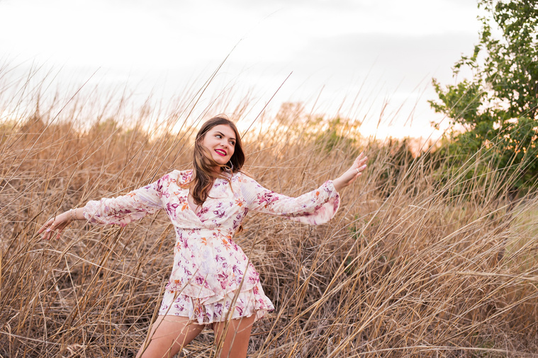 high school senior girl dressed in boho style stands in tall golden grass with both arms out in a carefree way looking to the side at sunset in Oklahoma City, Oklahoma