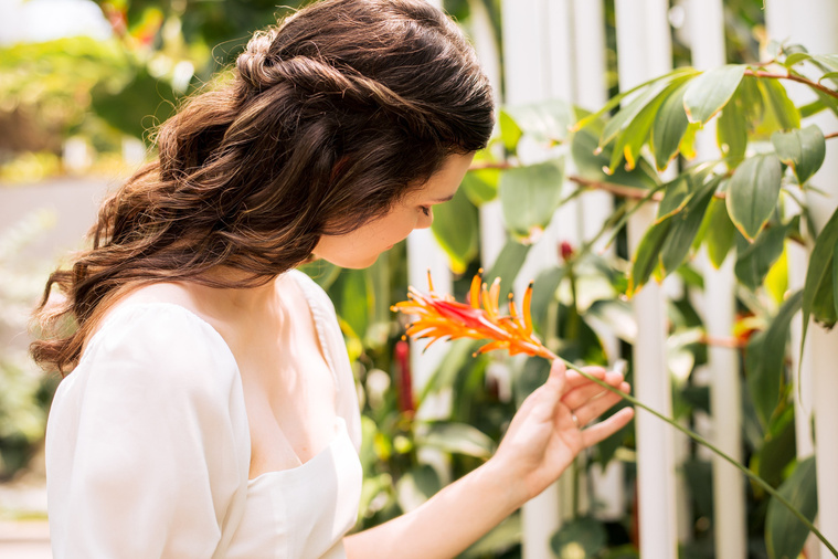 high school senior girl with long brown hair in a white blouse looks down at an orange tropical flower inside the Crystal Bridge at Myriad Gardens in Oklahoma City 