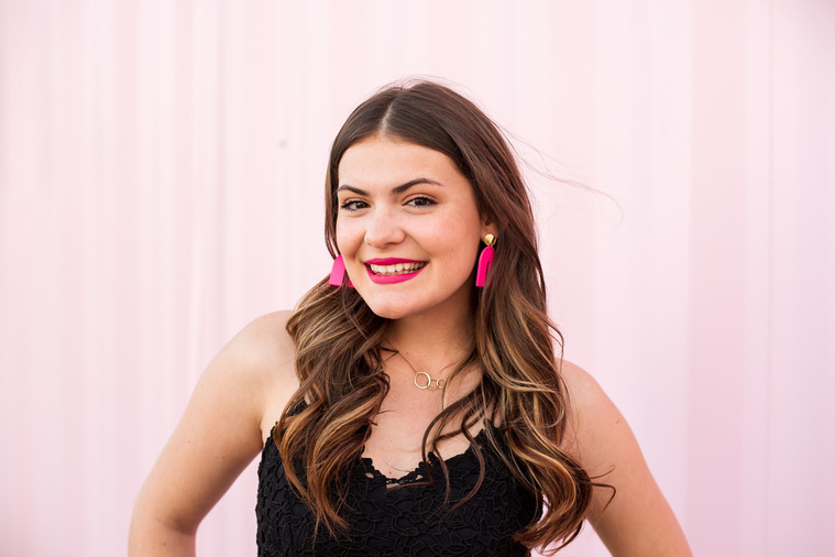 high school senior girl with long brown hair, black dress and pink earrings standing in front of a light pink wall in Automobile Alley in Oklahoma City, Oklahoma