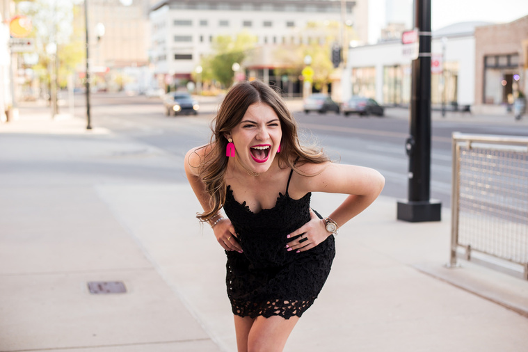 high school senior girl wearing a black dress and pink earrings has her hands on her hips and is laughing hard while she stand on the sidewalk in Automobile Alley in Oklahoma City, Oklahoma