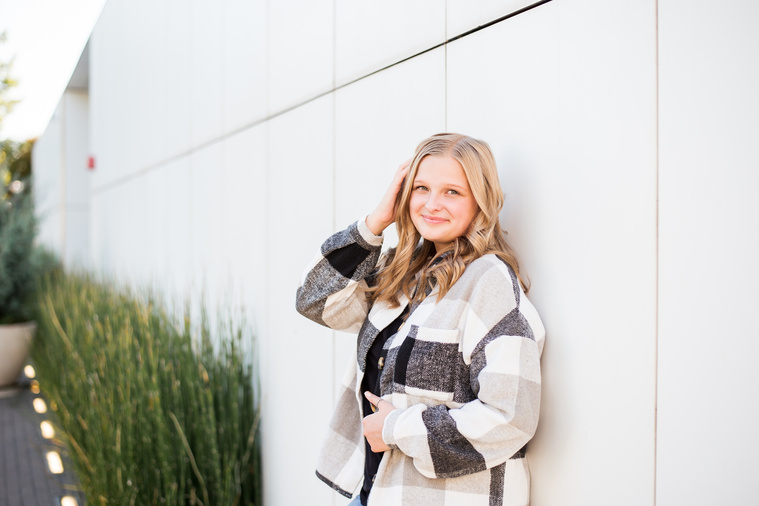blond high school senior girl wearing a black and white jacket standing in front of white wall at Myriad Gardens in downtown Oklahoma City