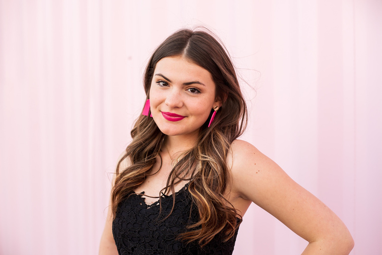 high school senior girl with long brown hair wearing a black dress and pink earrings standing in front of a pink wall and smiling with hand on hip in Automobile Alley in Oklahoma City, Oklahoma