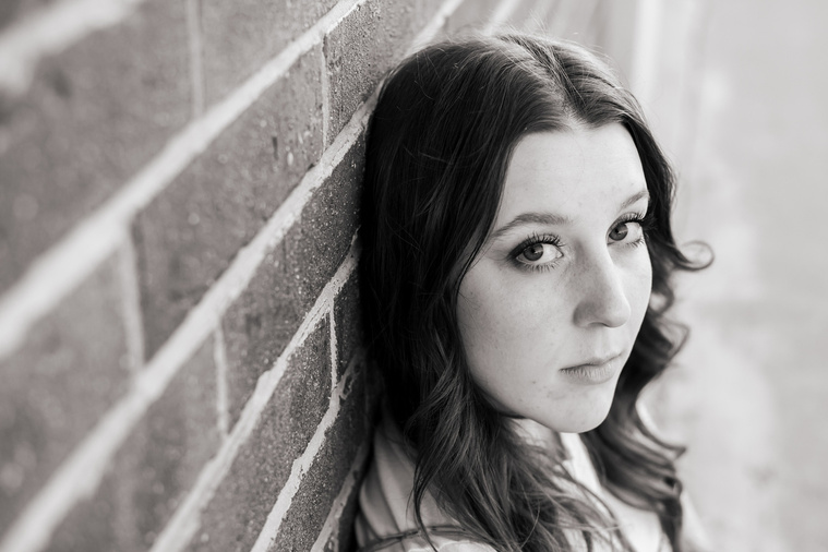 black and white landscape portrait of a senior girl sitting against a brick wall with a serious expression 