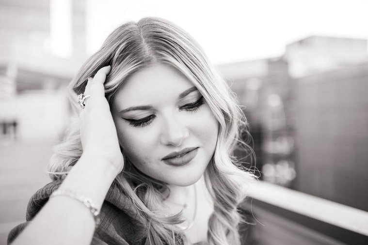 black and white close up image of a blond high school senior girl with one hand in her hair looking down softly smiling on top of the devon tower parking garage in okc