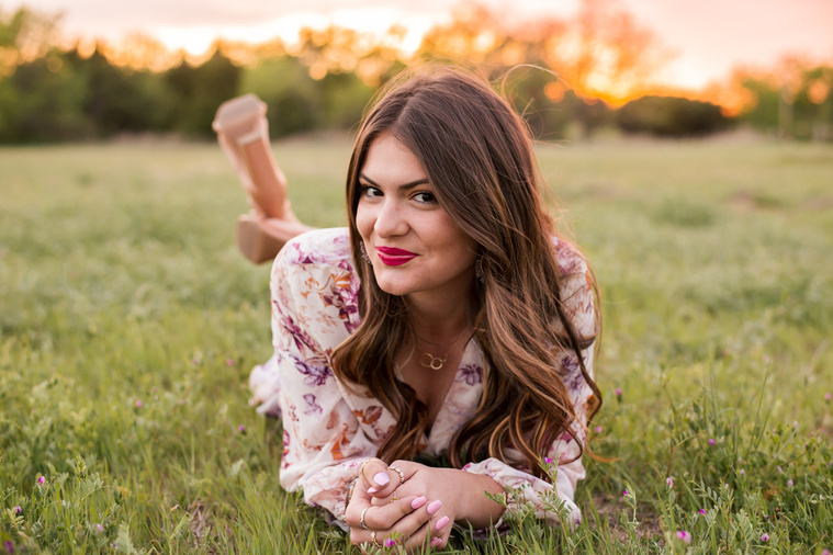high school senior girl dressed in boho style lays on her stomach and smiles to the side  in a field with trees and a sunset behind her in Oklahoma City, Oklahoma