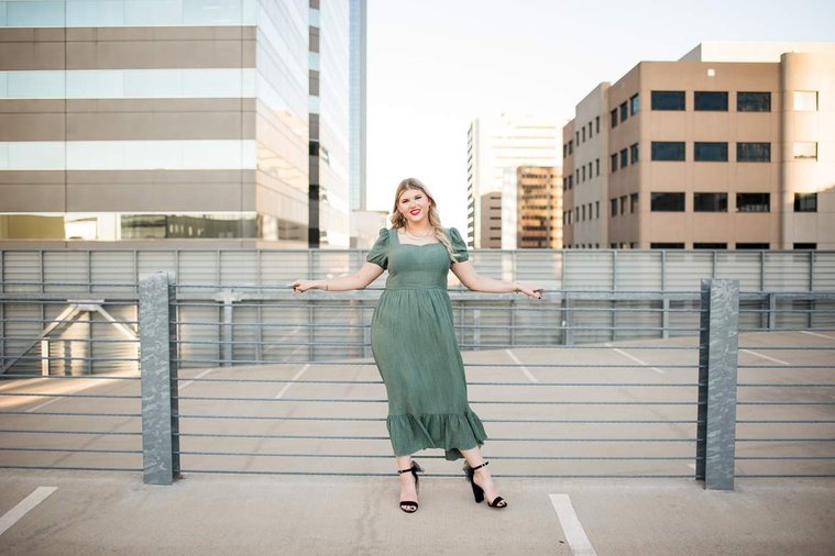 blond high school senior girl stands with both arms out and resting on wire fence in a green dress on top of the devon tower parking garage in okc