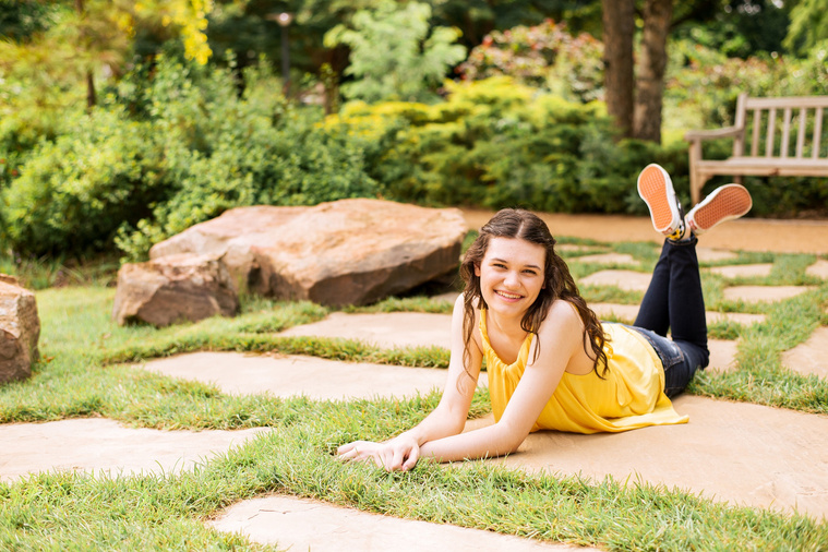 high school senior girl with long brown hair wearing a yellow top and jeans leans lays on her stomach with ankles crossed on a rock path with trees behind her and smiles at Myriad Gardens in Oklahoma City