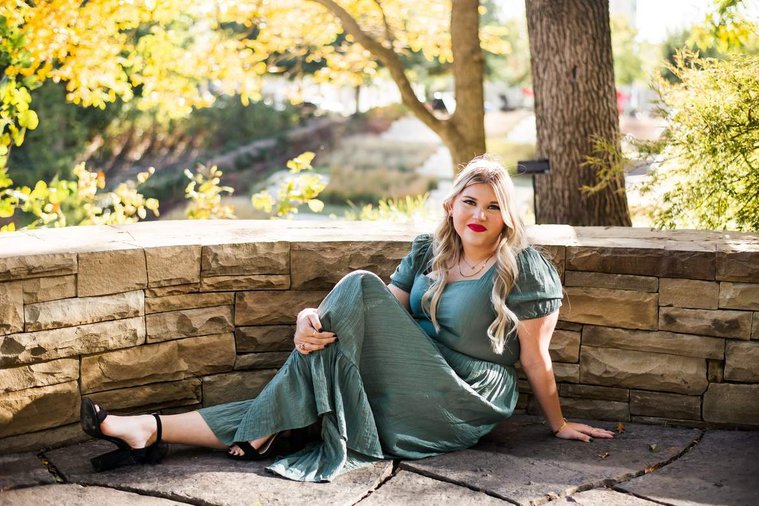 blond high school senior girl wearing a green dress is sitting on ground with stone wall behind her with one leg up and smiling at Myriad Gardens in downtown Oklahoma City for senior portraits