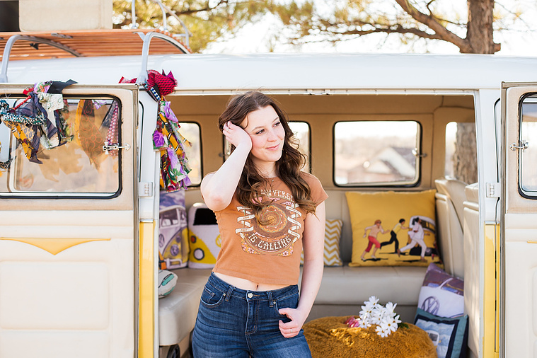 high school senior girl with brown hair wearing a t-shirt and bell bottoms stands in front of open side doors of a yellow and white VW bus in a yard in Tuttle Oklahoma with one hand in her pocket and the other in her hair