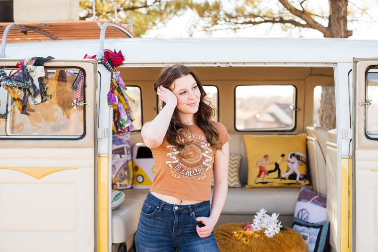 high school senior girl with brown hair wearing a t-shirt and bell bottoms stands in front of open side doors of a yellow and white VW bus in a yard in Tuttle Oklahoma with one hand in her pocket and the other in her hair