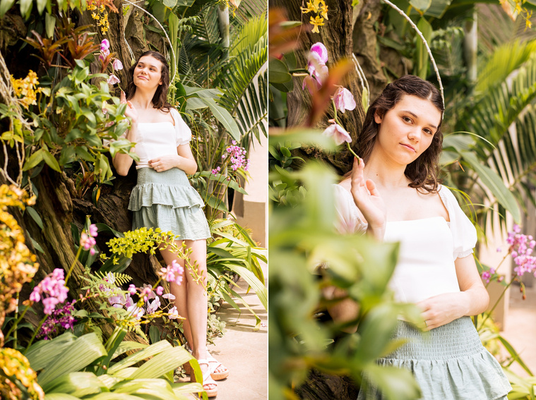 two photos of a high school senior girl with long brown hair in a white top and green skirt stands among a tropical plant wall with purple flowers inside the Crystal Bridge at Myriad Gardens in Oklahoma City 