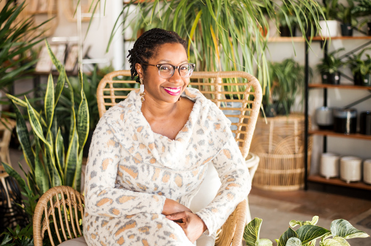  a professional woman sits in a wicker chair and smiles in front of plants at a plant shop in Oklahoma City Oklahoma