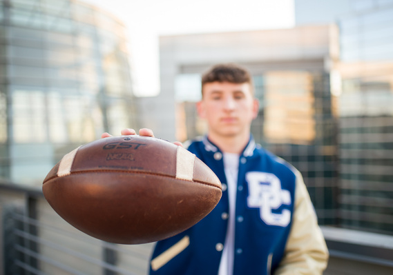 high school senior boy wearing letterman jacket holds a football close to camera, football is in focus and he has a game face on. standing on the rooftop of a parking garage in downtown OKC with the Devon Tower behind him