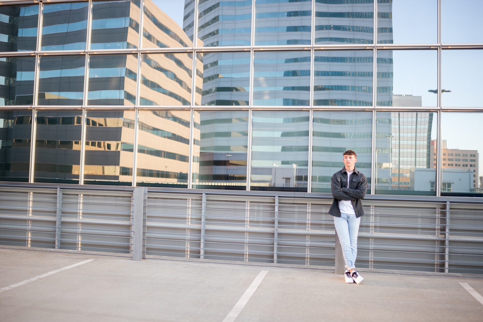 senior guy wearing gray jacket and light jeans with sneakers leans against a parking garage rooftop wall with the reflective windows of the Devon Tower in downtown OKC behind him