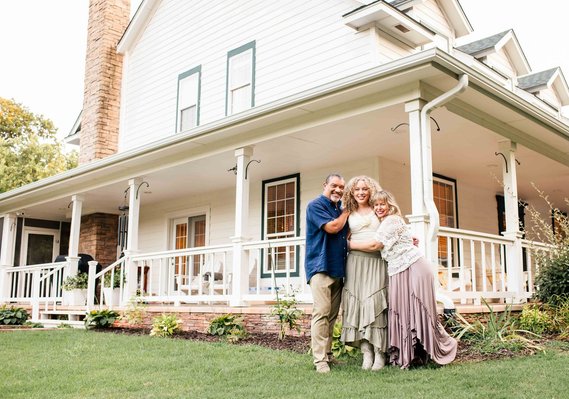 dad, mom, and teen daughter stand together hugging and smiling in front of their white country home with a wraparound porch near Oklahoma City, Oklahoma.