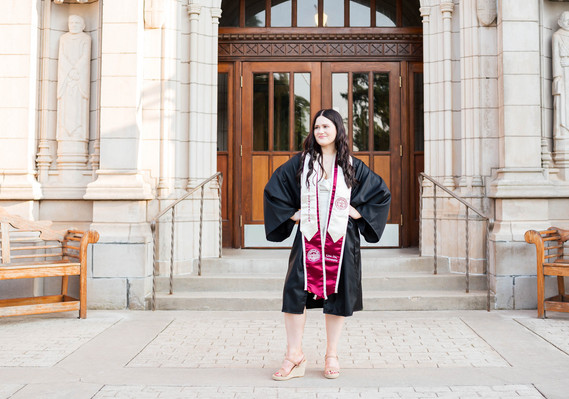 OU college senior girl with long dark hair stands in front of entry doors on campus in Norman in her graduation gown and stole, with hands on her hips and smiling off to the side