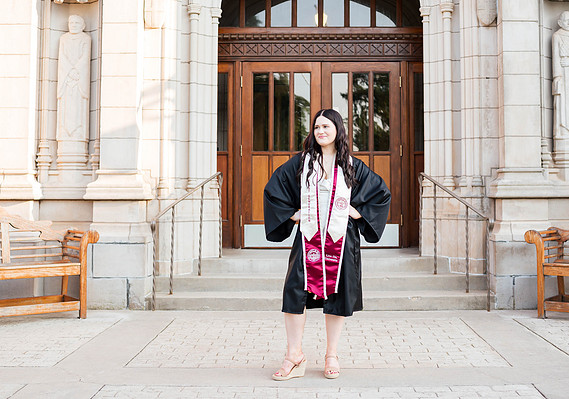 OU college senior girl with long dark hair stands in front of entry doors on campus in Norman in her graduation gown and stole, with hands on her hips and smiling off to the side