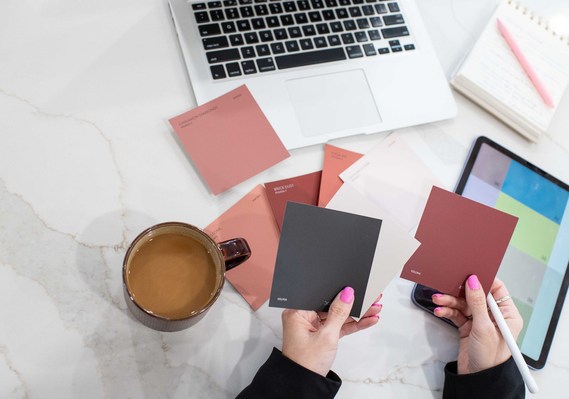 overhead shot of a designer comparing color swatches in each hand with a color pixel grid on her tablet and laptop, notebook and coffee surrounding her. 