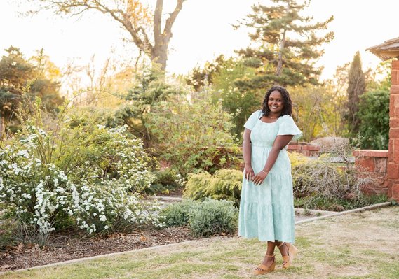 black senior girl wearing a green dress smiles prettily while she stands in front of pretty flowers and trees at Will Rogers Gardens in Oklahoma City