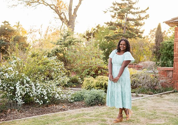 black senior girl wearing a green dress smiles prettily while she stands in front of pretty flowers and trees at Will Rogers Gardens in Oklahoma City