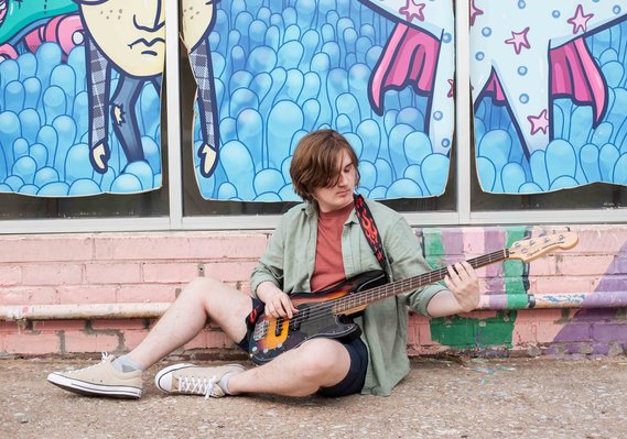 senior boy in button down and shorts sits on the sidewalk holding his bass guitar in his lap and looking at it in front of a colorful wall mural in midtown Oklahoma City