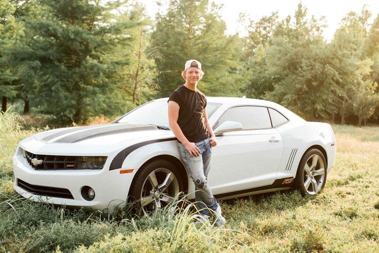 high school senior boy wearing a backwards baseball cap, black shirt and blue jeans leans against his Chevy Camaro parked in a green field with trees behind him in Tuttle, Oklahoma
