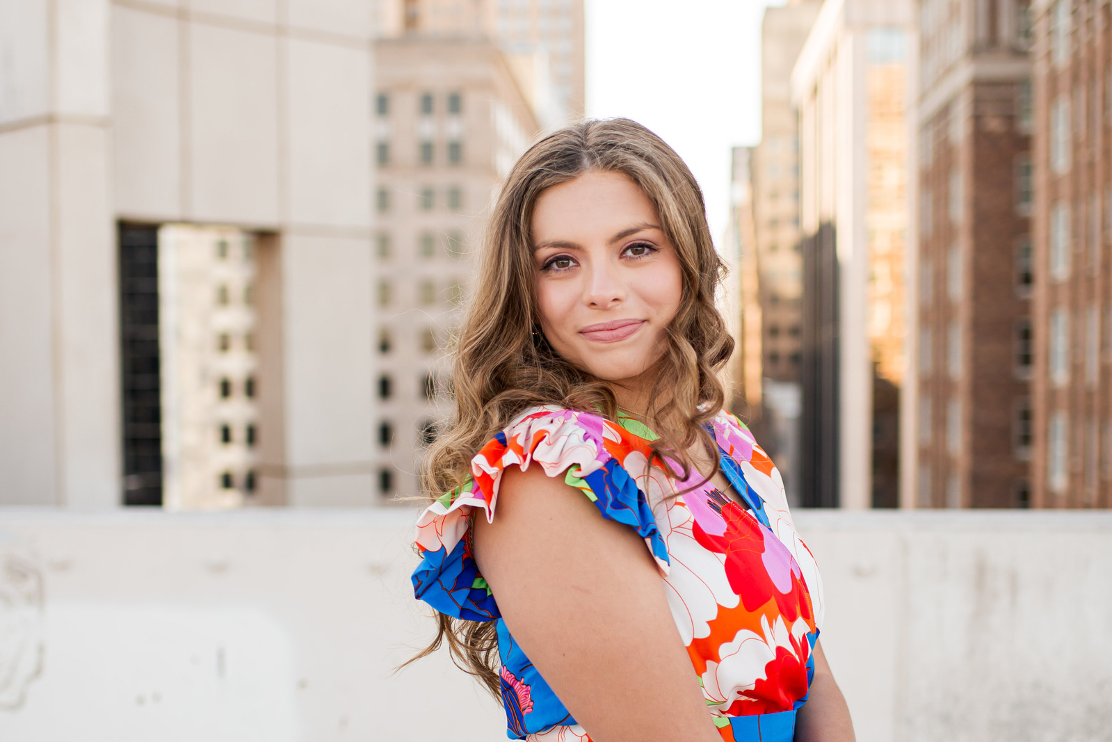 senior girl with long curly hair wearing a patterned colorful dress smiles sweetly over her shoulder standing on the rooftop of the Santa Fe parking garage in OKC