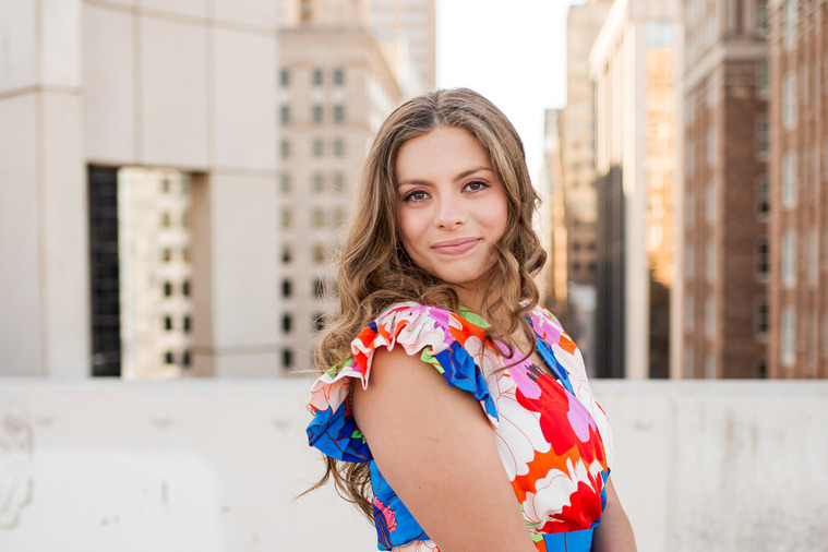 pretty senior girl wearing a bright floral dress smiles softly over her shoulder standing on the Santa Fe parking garage rooftop in OKC