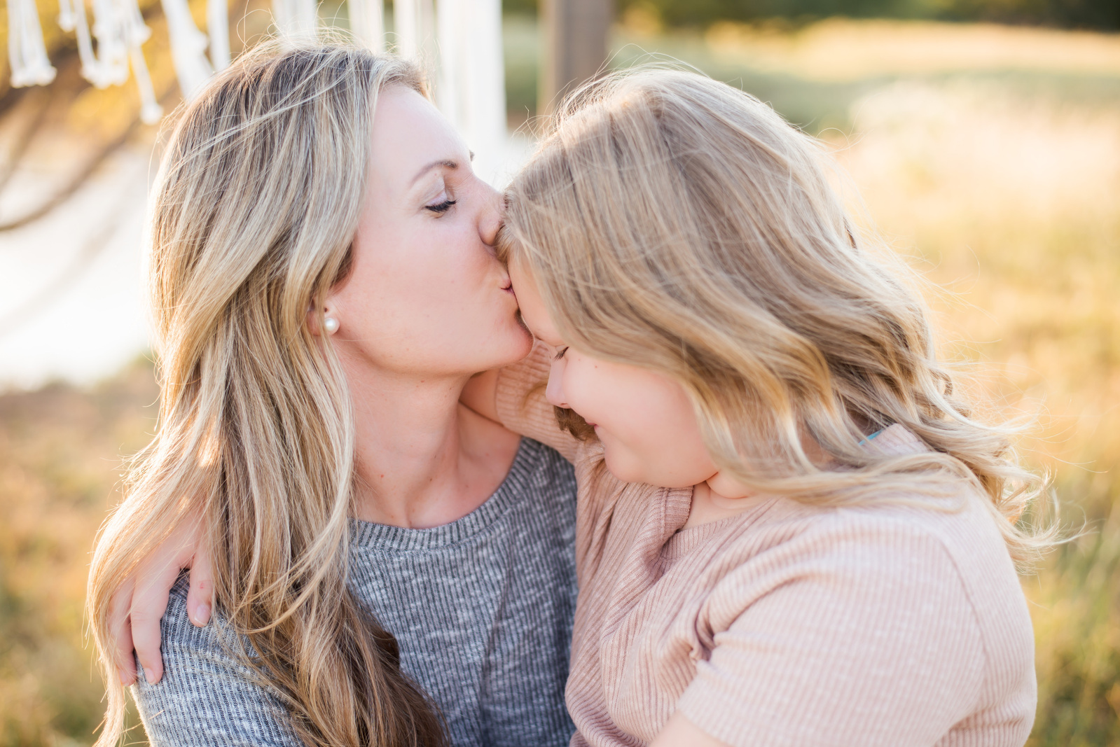 a mom with long blond hair kisses the forehead of her 10 year old daughter with blond hair with golden grass and a pond behind them in a field in central Oklahoma