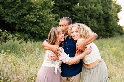 a dad hugs his daughter and wife close to him and kisses his wife's forehead as they laugh together in an open field with trees in the country near Norman Oklahoma