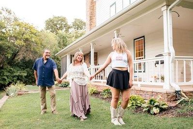 parents and an older teen daughter hold hands and walk together in front of their house and porch while she looks back at them wearing her OU college gear
