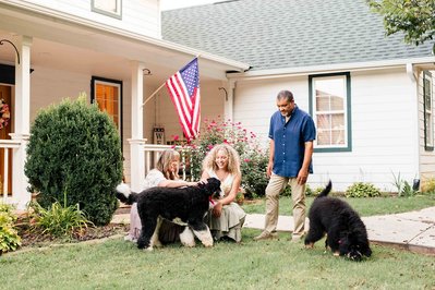 a family of 3 pose candidly in front of their home and porch with their two energetic dogs who they are trying to get to sit for a picture
