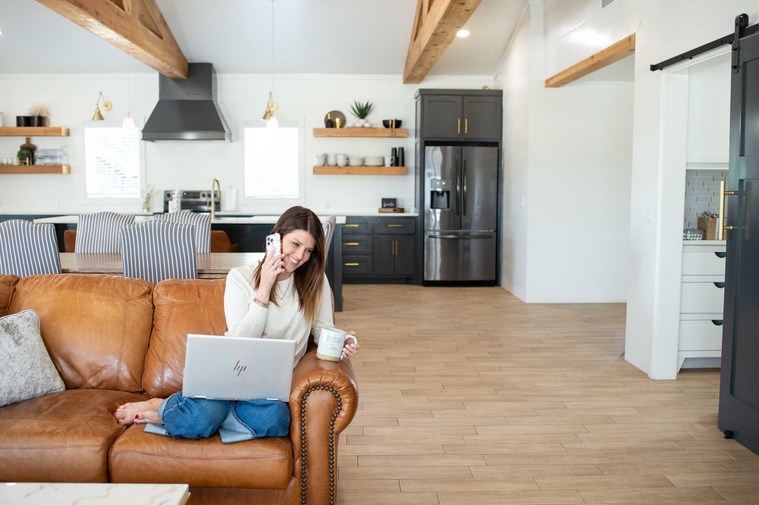 attractive female realtor sits cozily on a couch at home with open laptop, on the phone with a buyer, holding a mug of coffee at a brand session in Newcastle Oklahoma.