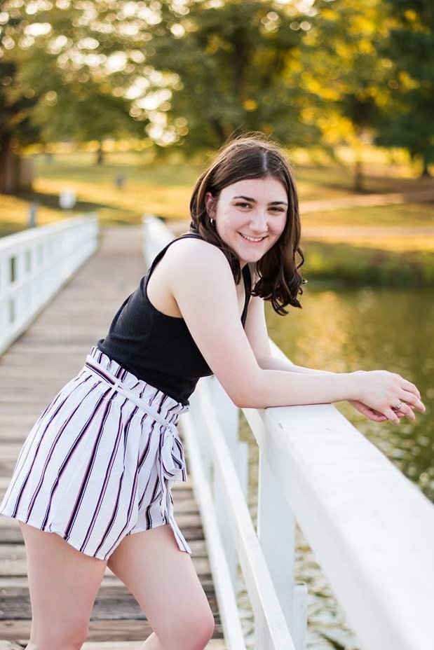 A high school senior girl stands on a white bridge and smiles in front of the trees and pond in Shannon Springs Park in Chickasha, Oklahoma 