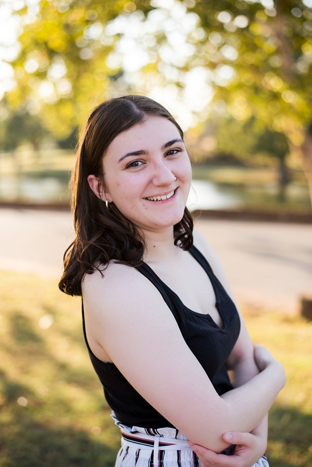 A high school senior girl stands and smiles in front of the trees and pond in Shannon Springs Park in Chickasha, Oklahoma 