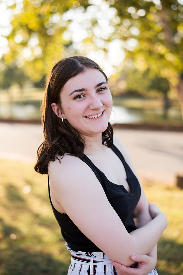 A high school senior girl stands and smiles in front of the trees and pond in Shannon Springs Park in Chickasha, Oklahoma 