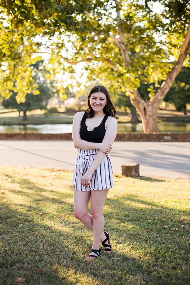 A high school senior girl stands and smiles in front of the trees and pond in Shannon Springs Park in Chickasha, Oklahoma 