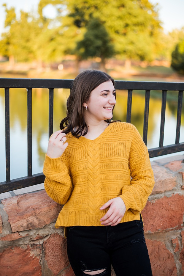 A high school senior girl stands and smiles on a stone bridge in front of the trees and pond in Shannon Springs Park in Chickasha, Oklahoma 
