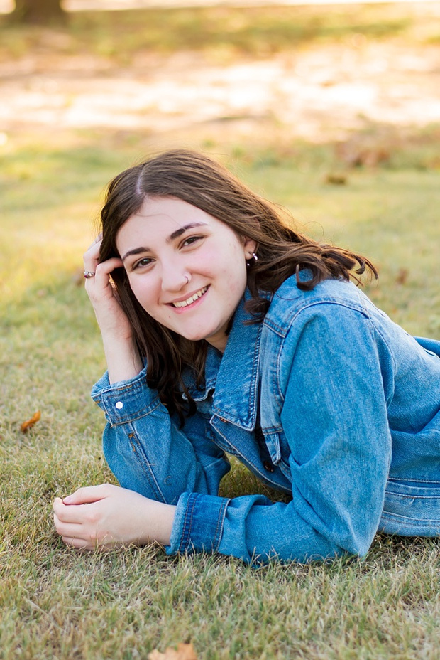 A high school senior girl laying in the grass and smiles in front of the trees and pond in Shannon Springs Park in Chickasha, Oklahoma 