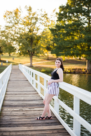 A high school senior girl stands on a walking bridge and smiles in front of the trees and pond in Shannon Springs Park in Chickasha, Oklahoma 
