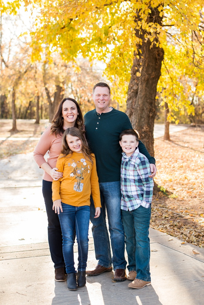 a mom, dad, young son and daughter stand together on a sidewalk in a park in Moore, Oklahoma for a family photo session with a tree with bright golden leaves behind them