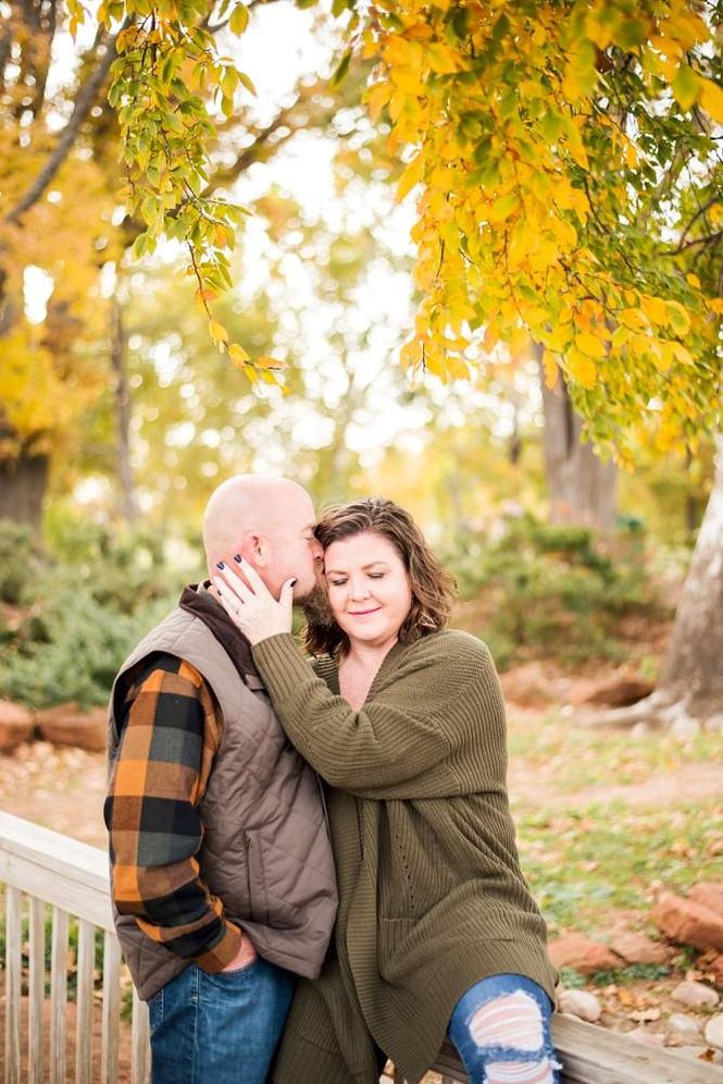 A husband and wife stand on a footbridge and snuggle together in a quiet moment with golden fall colored leaves hanging above them at Will Rogers Gardens in Oklahoma City, Oklahoma