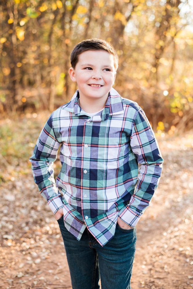 a cute young boy stands with both hands in his pockets and smiles off camera for a portrait at a family photo session in Moore, Oklahoma in the woods in fall