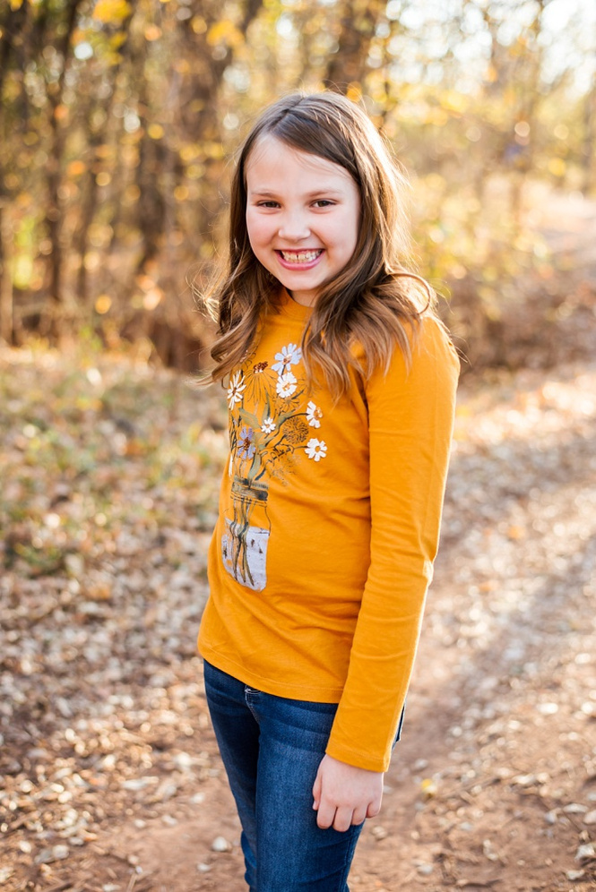a cute young girl stands and smiles at the camera for a portrait in the woods with fall color around her in moore, oklahoma at a family photo session