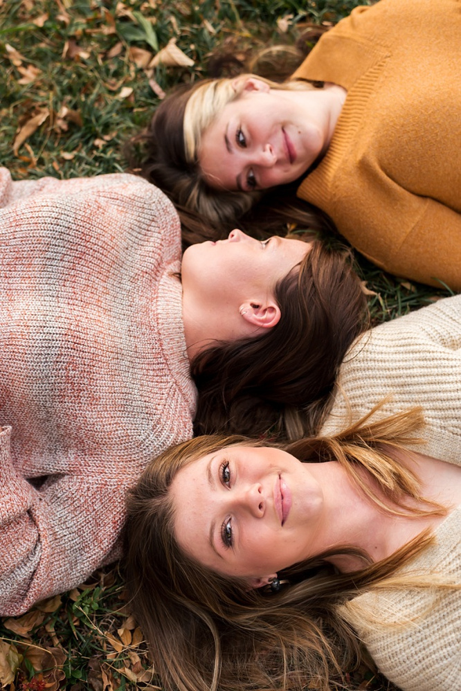 3 teen girls lay on their backs with their faces close to one another in a line. Two look at each other, the third is looking and smiling at the camera,  at Will Rogers Gardens in Oklahoma City, Oklahoma